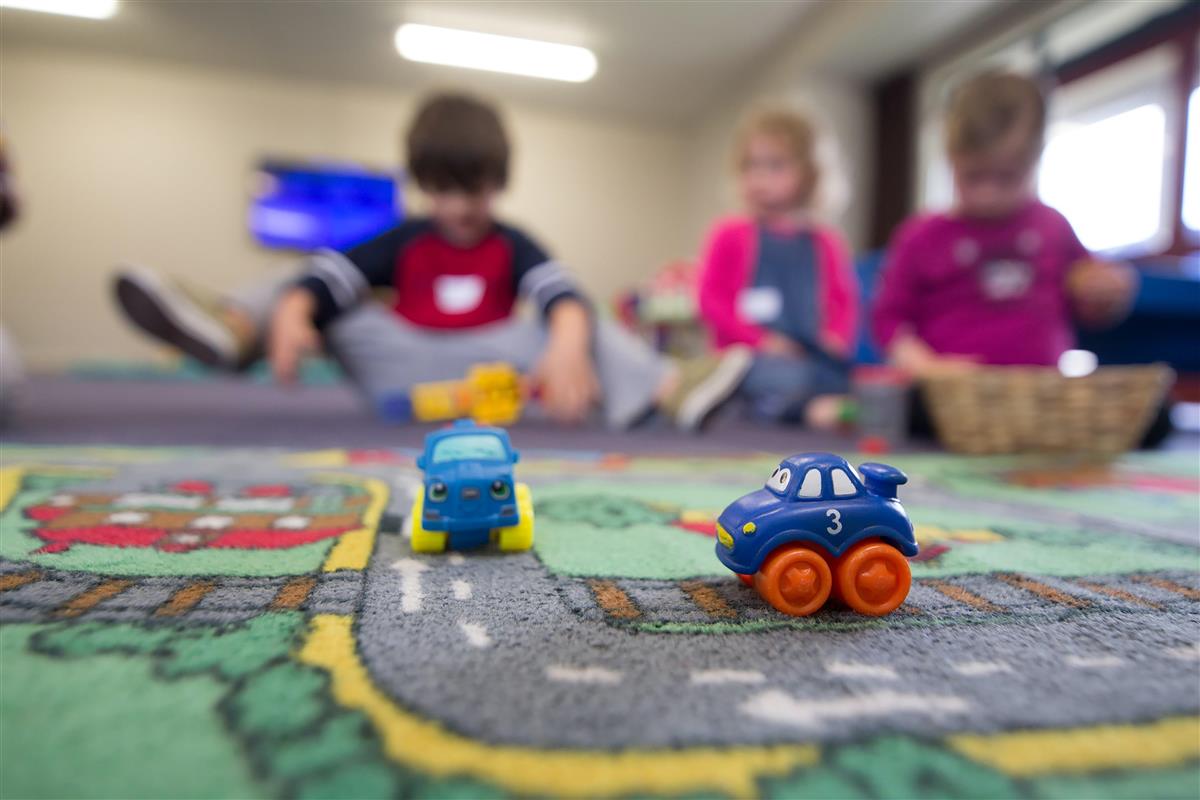 Barn i en barnehage i bakgrunnen, lekebiler i fokus i front av bildet. Foto. - Klikk for stort bilde